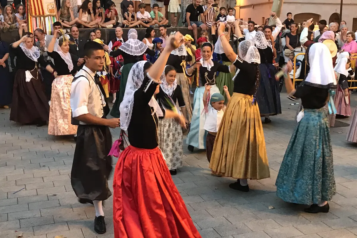 Dancers in traditional dress, women in white lace headscarves and a long red skirt, dancing in a paved square with spectators behind