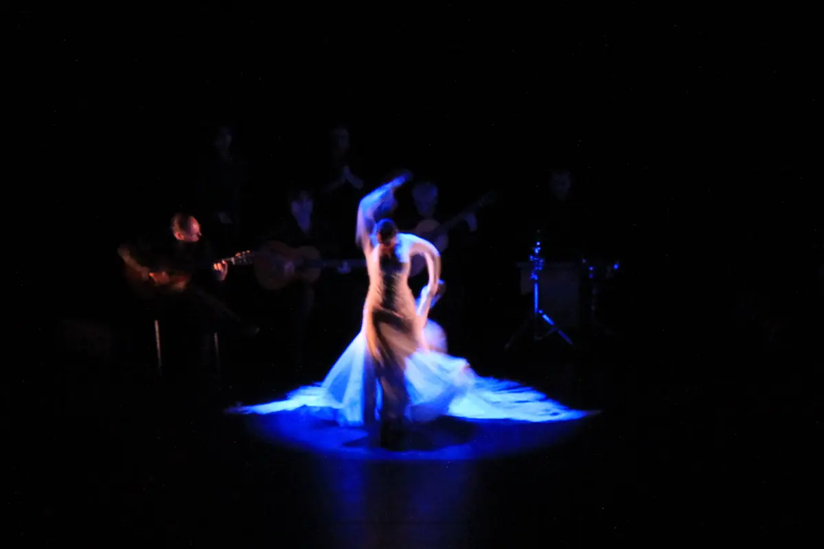 A flamenco dancer in a flowing skirt lit in blue on a dark stage, with guitarists in the shadows behind her