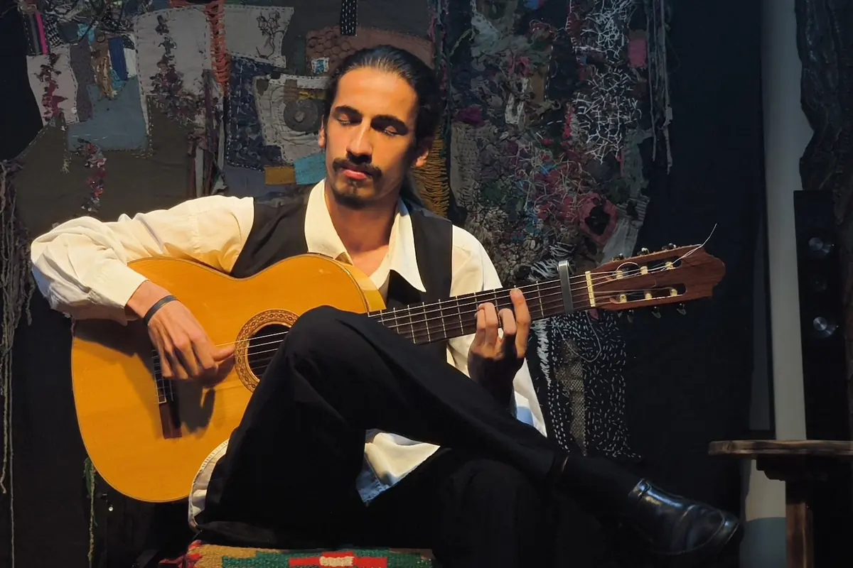 A seated guitarist in a waistcoat playing a Spanish guitar in front of a patterned textile backdrop